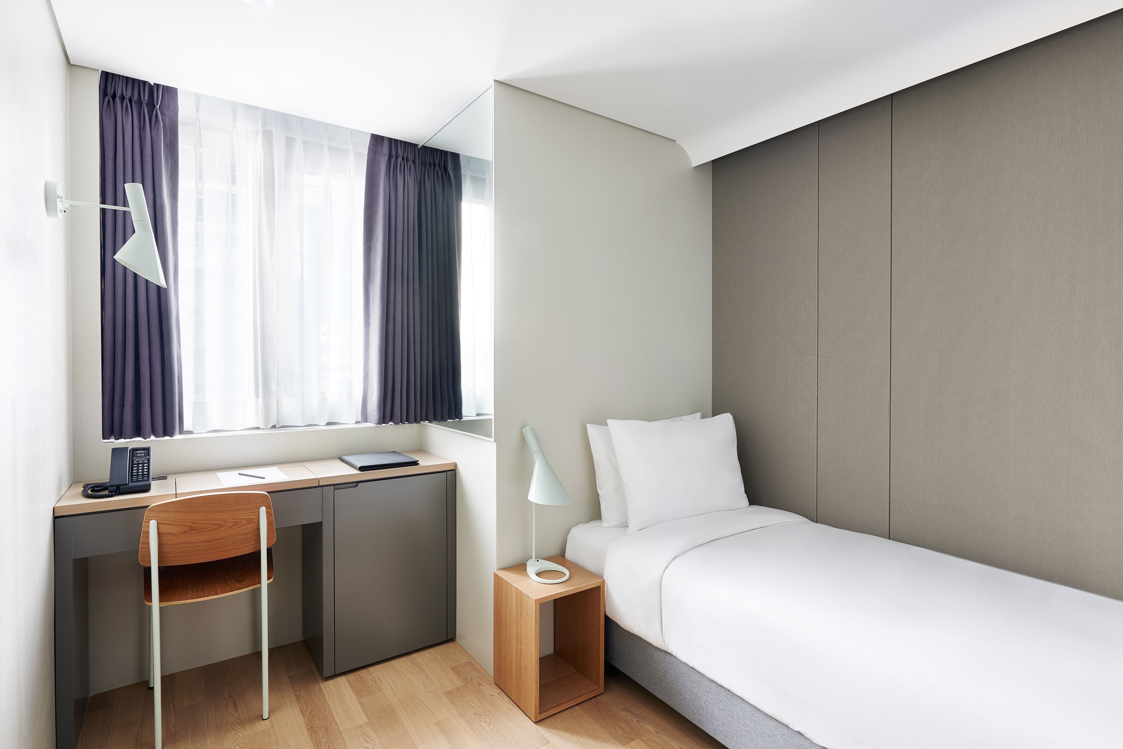 A modern hotel room with a desk, chair, bed, bedside table, lamp, and a window with curtains.
