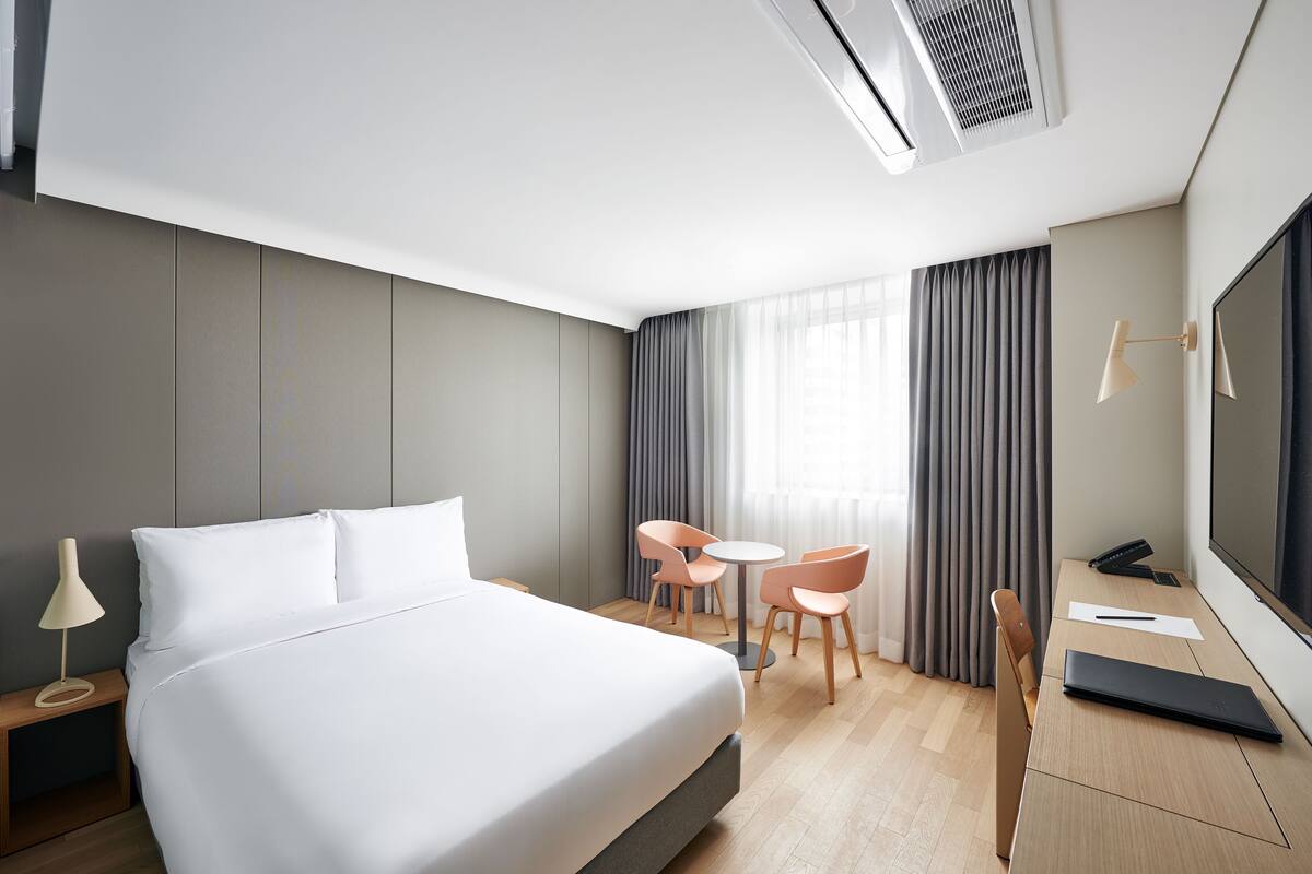 A modern hotel room with a large bed, a desk with a laptop, two chairs, a TV, and an air conditioning unit.