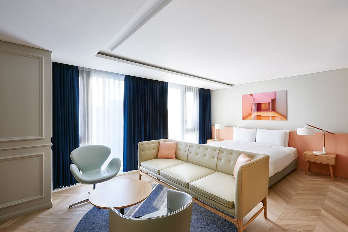 A modern hotel room with a sofa, a chair, a bed, a nightstand, and a lamp.