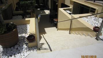 Un escalier recouvert de petits galets blancs, agrémenté de plantes en pot, mène à un espace extérieur abrité, aménagé avec une table et des chaises.