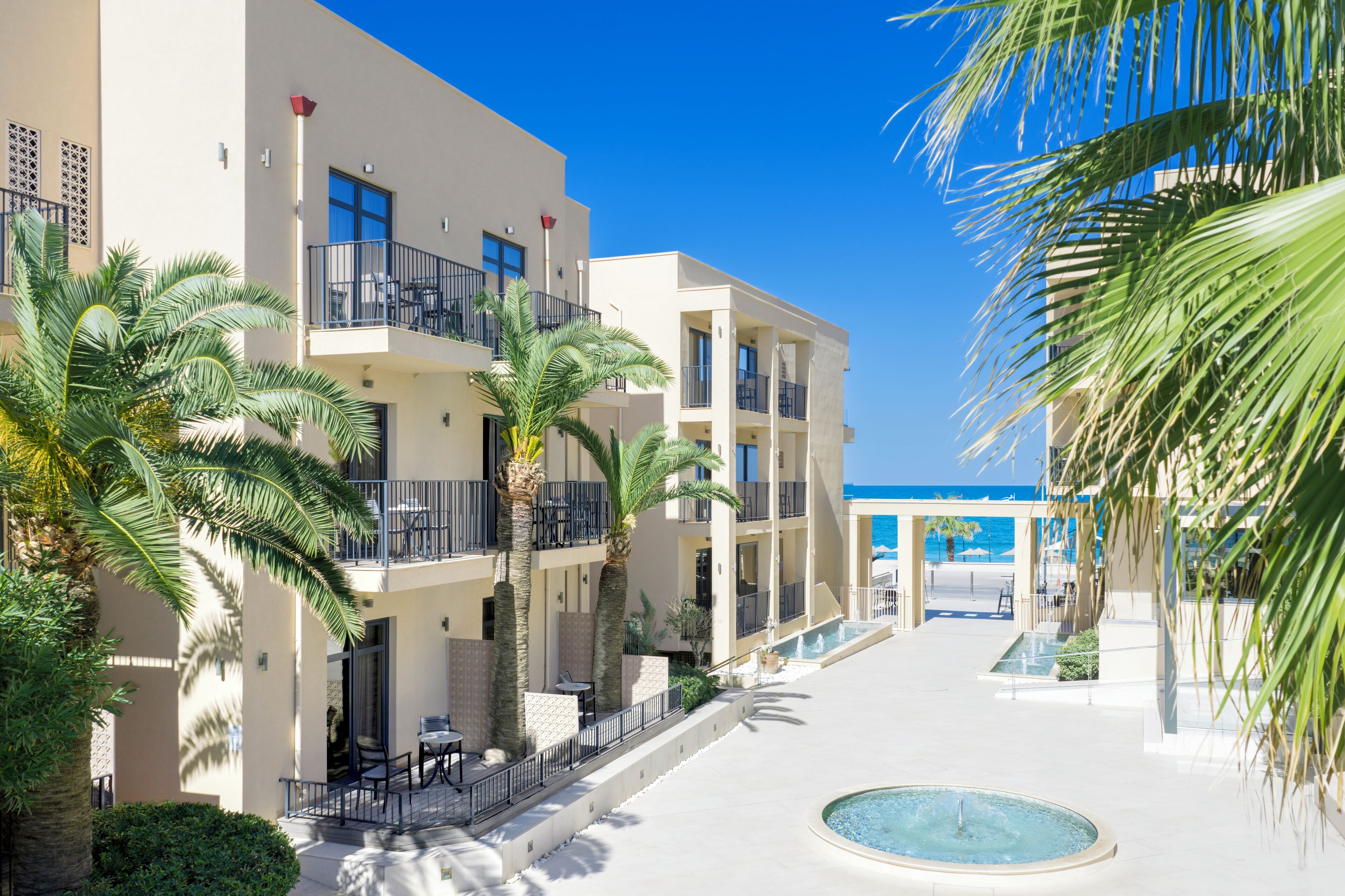 Un hotel moderno con varios balcones, palmeras y una zona de piscina central.
