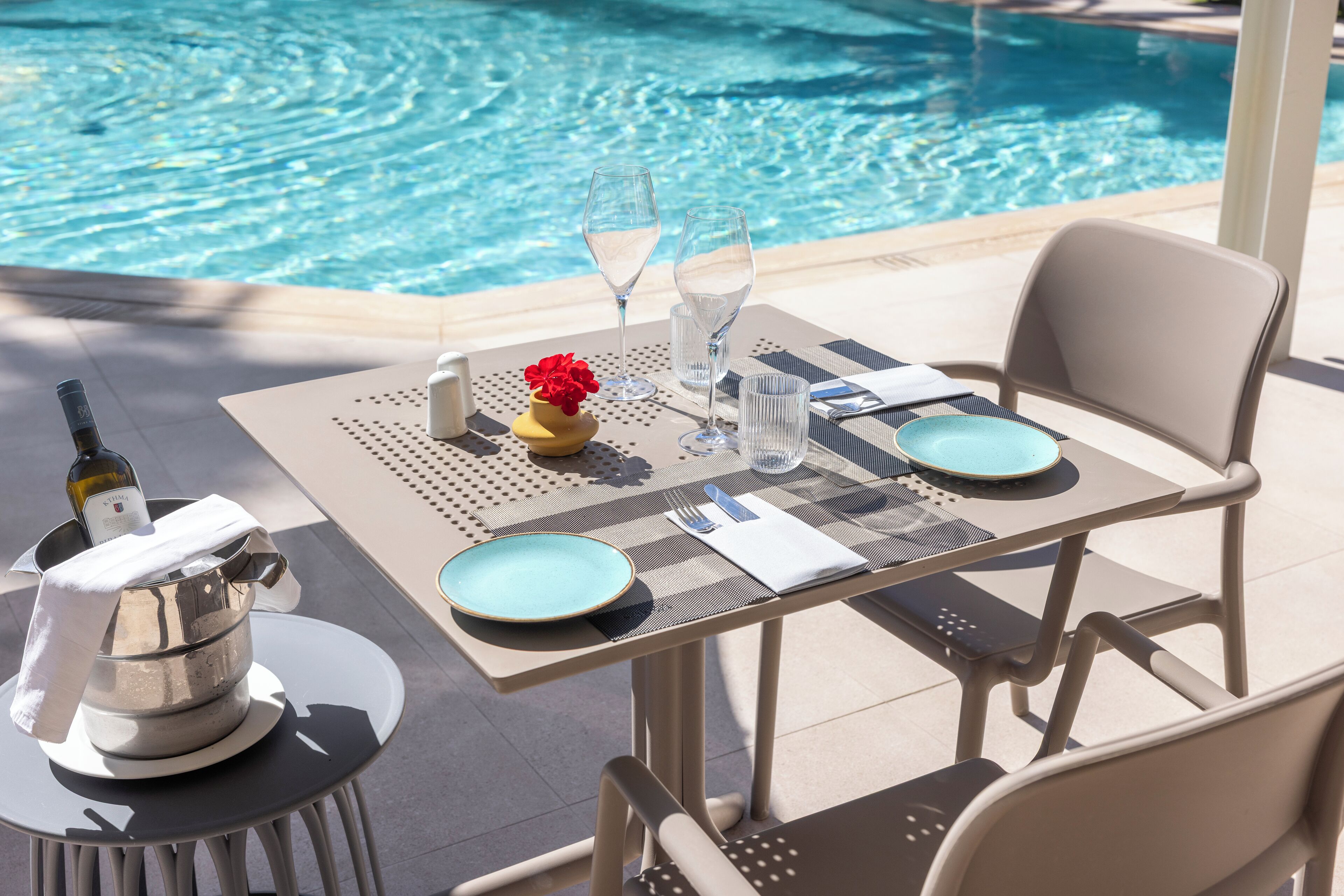 A table set for two by a pool with a bottle of champagne, two glasses, and a vase with red flowers.