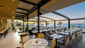 A restaurant with outdoor seating overlooking the sea, featuring tables and chairs arranged under a covered area.