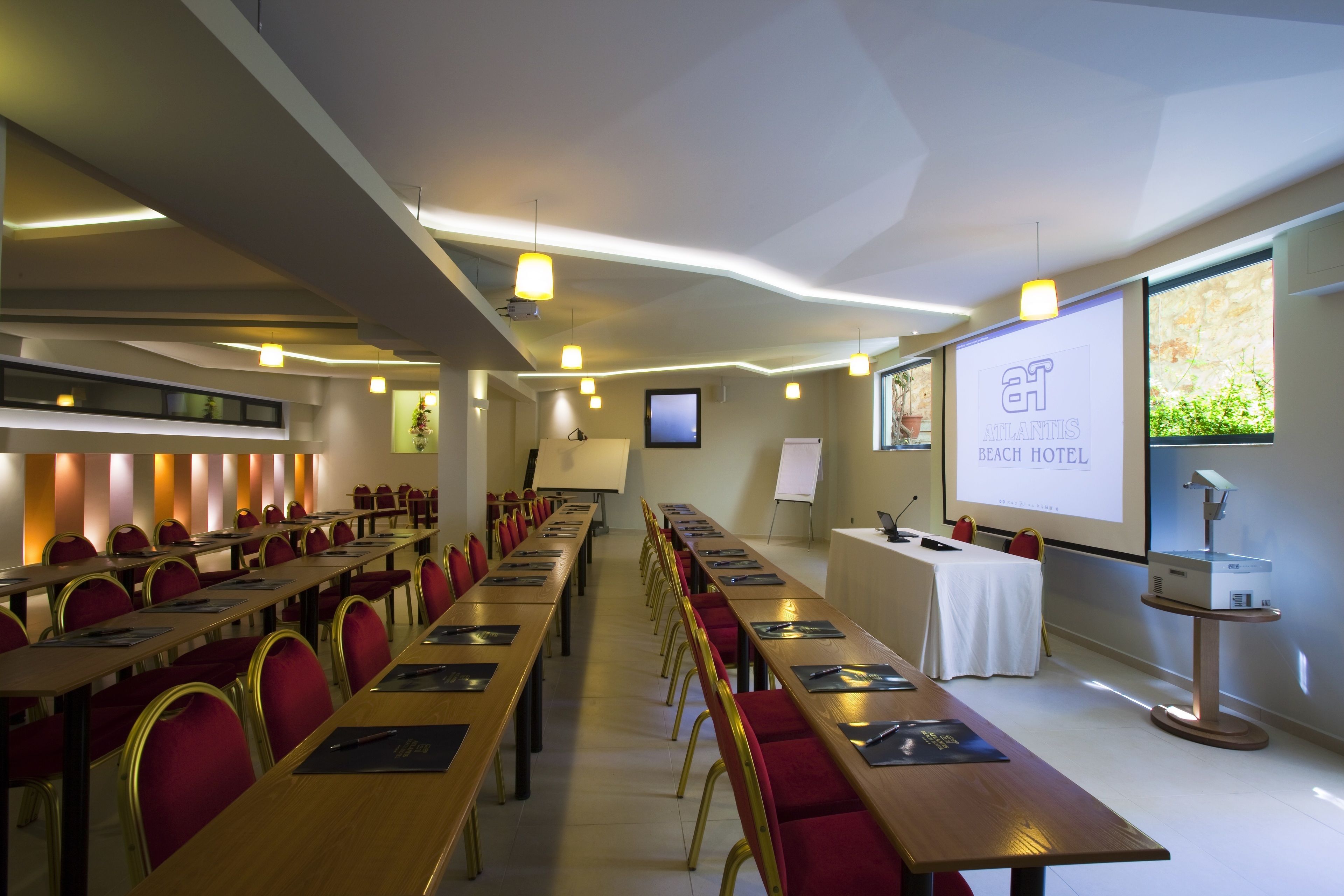 A conference room with long tables, red chairs, and a presentation screen displaying "AT ATLANTIS BEACH HOTEL".