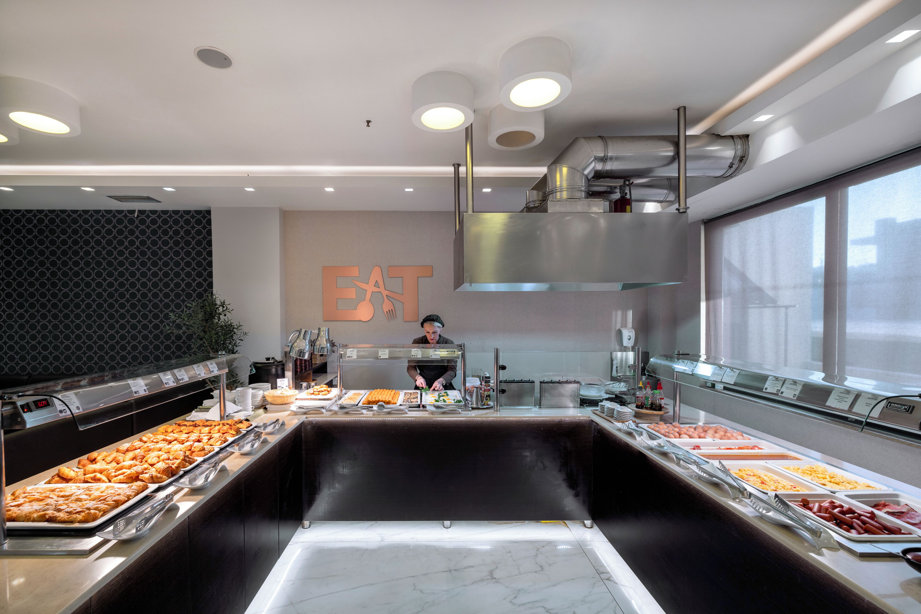 A buffet setup with various food items, a chef preparing food, and a large window providing natural light.
