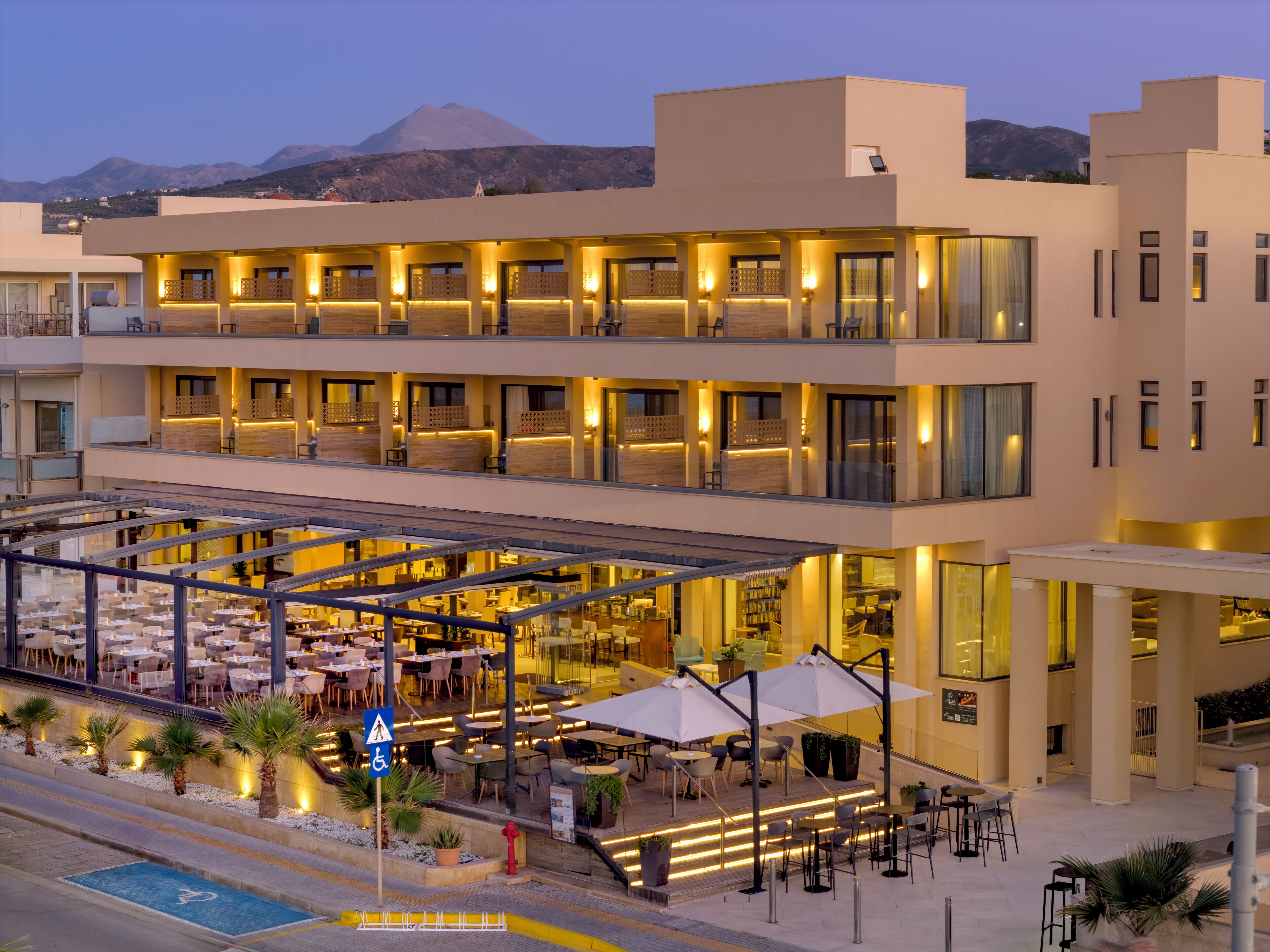 Un hotel moderno con varios pisos, asientos al aire libre y vistas a montañas al fondo.