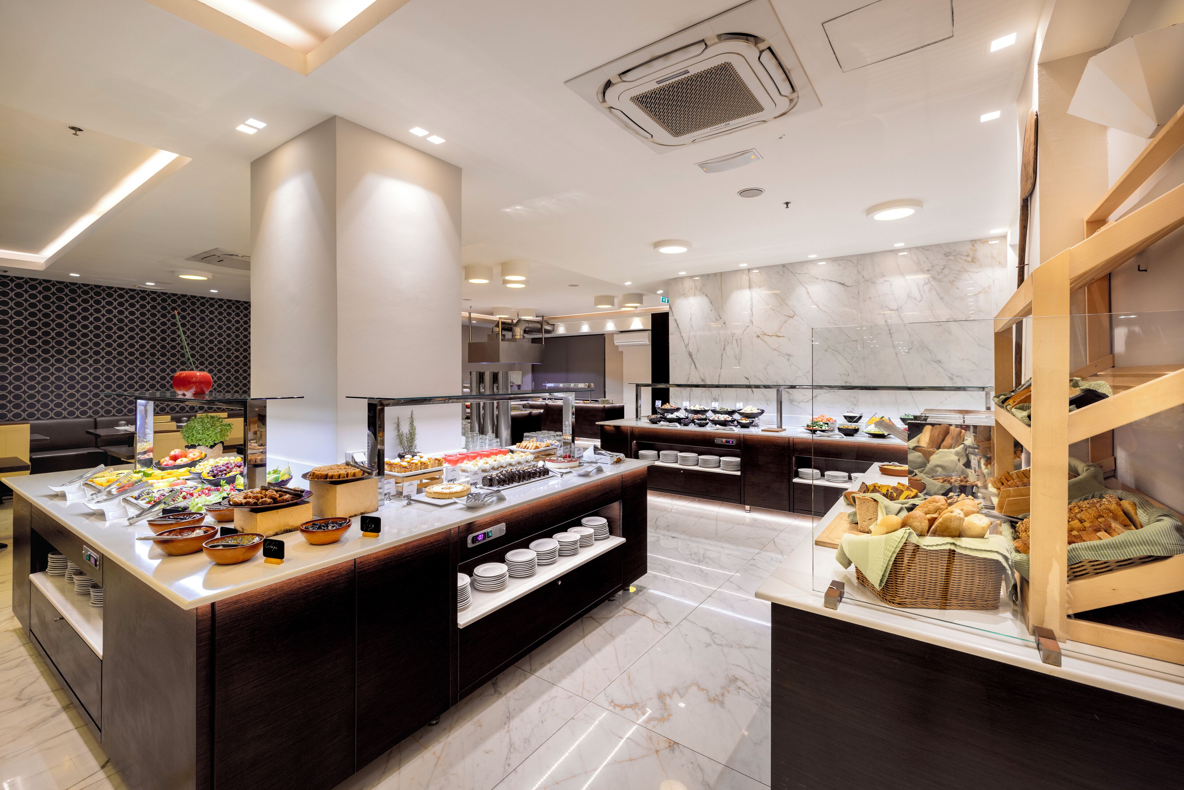 A modern hotel kitchen with marble countertops, a buffet area with various food items, and a wooden staircase.