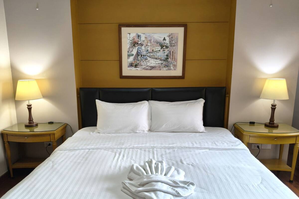A hotel room with a large bed, two bedside tables with lamps, a framed painting above the bed, and a folded towel on the bed.