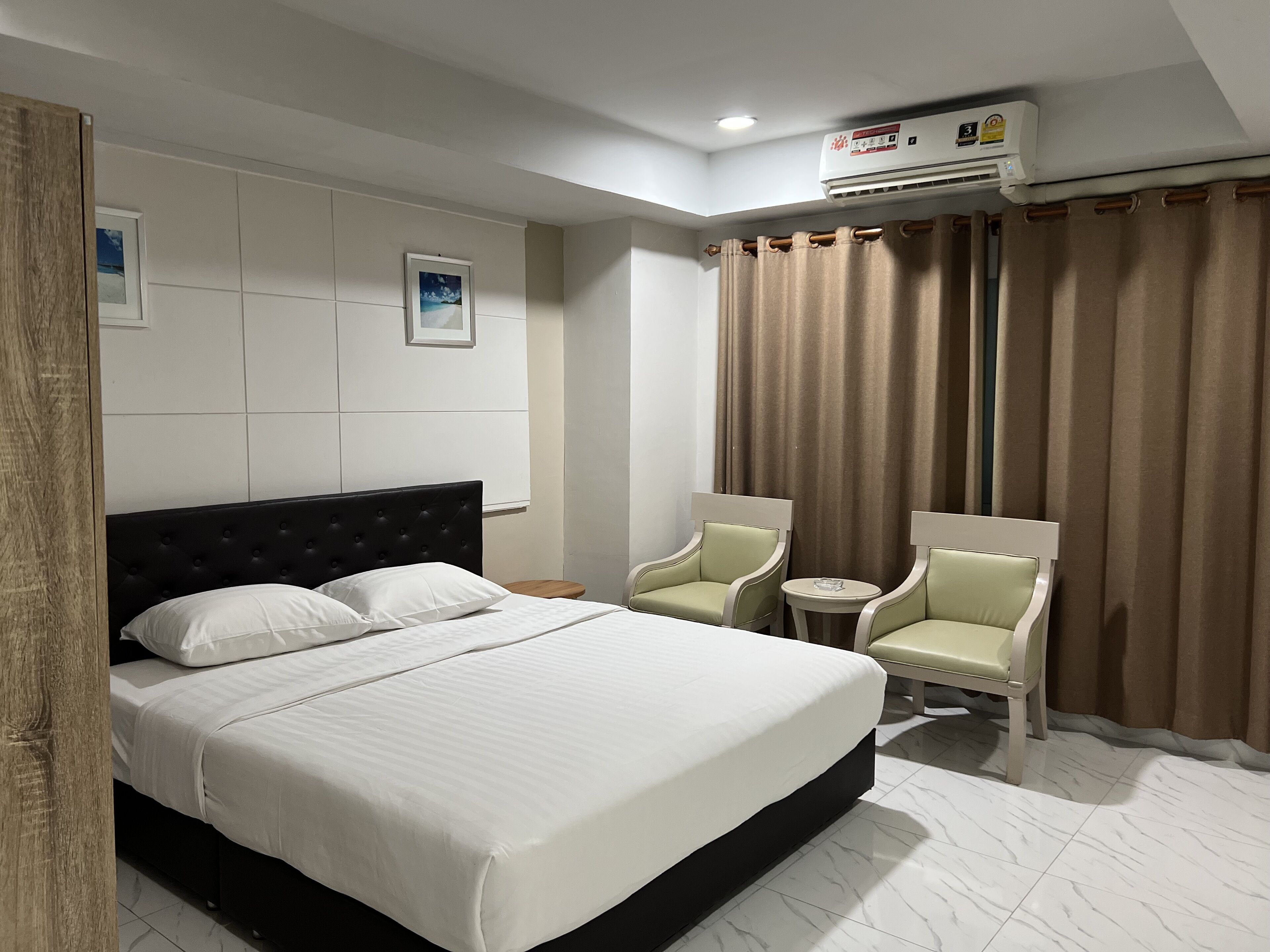A hotel room with a bed, two chairs, a small table, an air conditioning unit, and curtains.