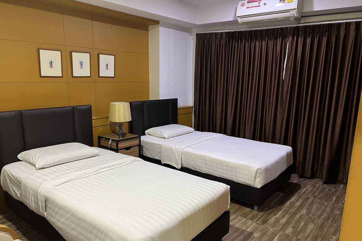 A hotel room with two beds, a nightstand with a lamp, a wall-mounted air conditioner, and a window with curtains.