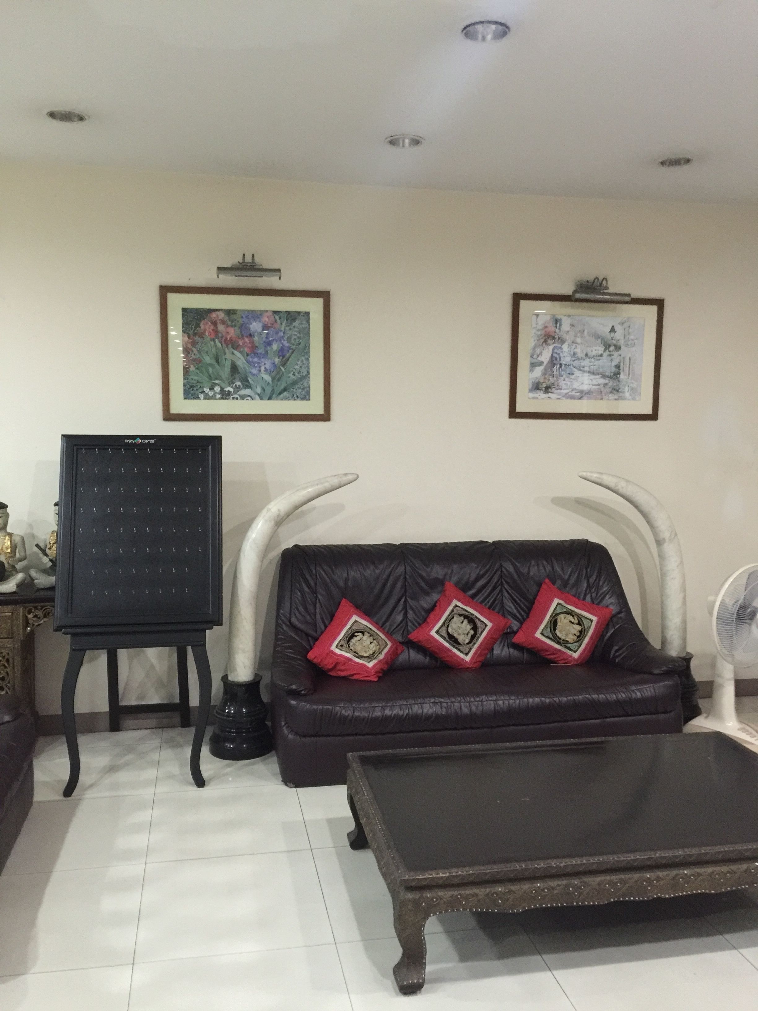 A living room with a black leather sofa, two framed pictures on the wall, a black easel, and a white fan.