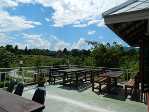 An outdoor patio with wooden tables and chairs, a railing, and a view of a green landscape with trees and a mountain in the distance. - Pai Iyara Resort (Pai)