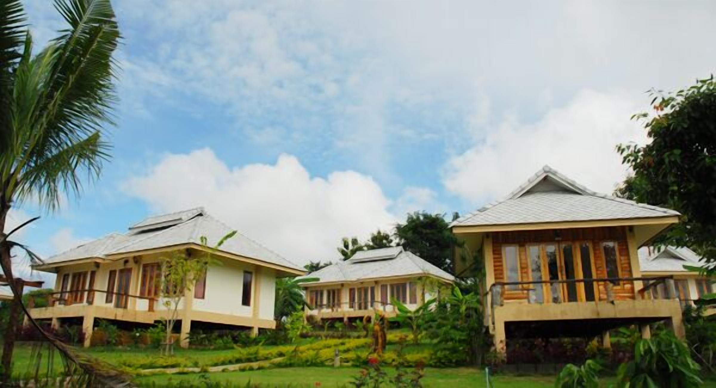 A row of three bungalow-style buildings with sloped roofs, surrounded by lush greenery and a clear sky.
