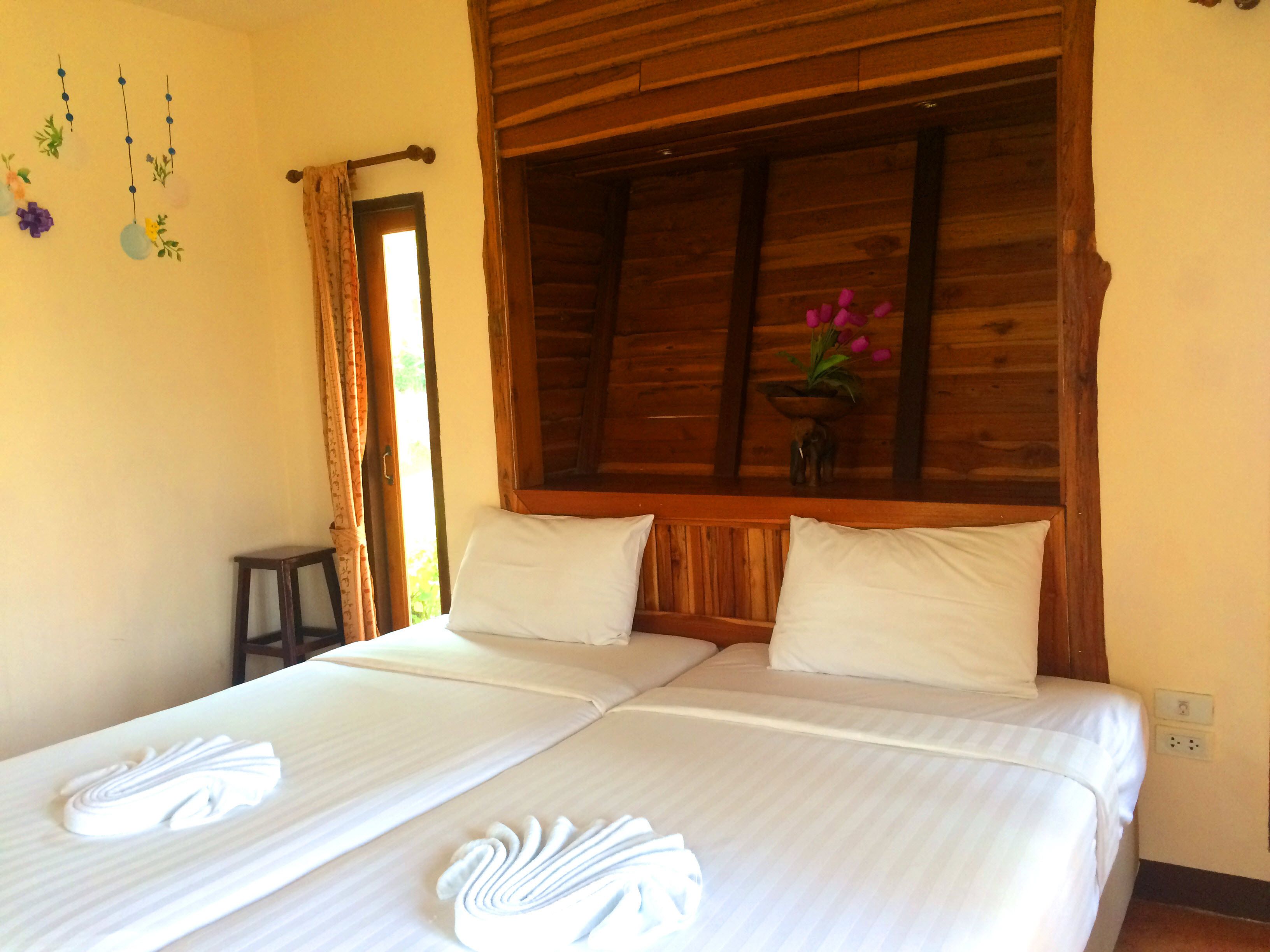 A double bed with white linens and towels, a wooden headboard, a small table, and a window with curtains.