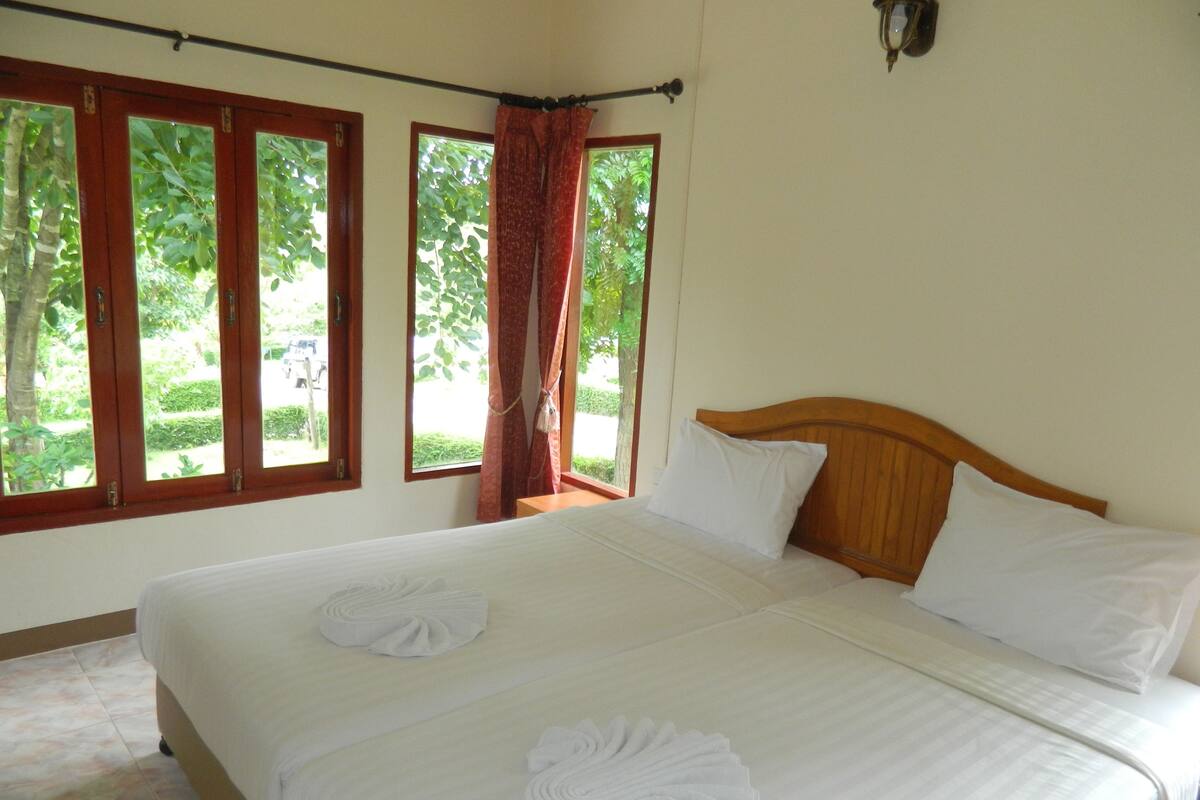 A hotel room with a large bed, two pillows, and a wooden headboard. There are three windows with curtains, and a view of greenery outside.