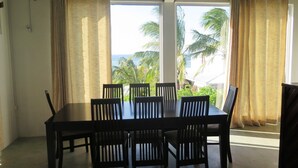 A dining area with a table and chairs, large windows with curtains, and a view of palm trees and the ocean. - Residence Foulsafat (Rodrigues Island)