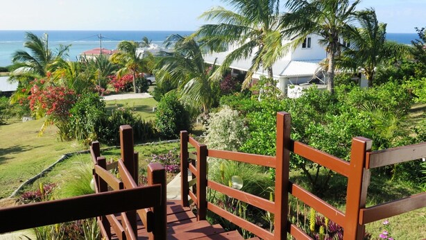 A wooden bridge leading to a white building with a view of the ocean and palm trees. - Residence Foulsafat (Rodrigues Island)