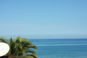 Visão nítida do oceano, com um barco ao longe, palmeiras e um guarda-sol branco.