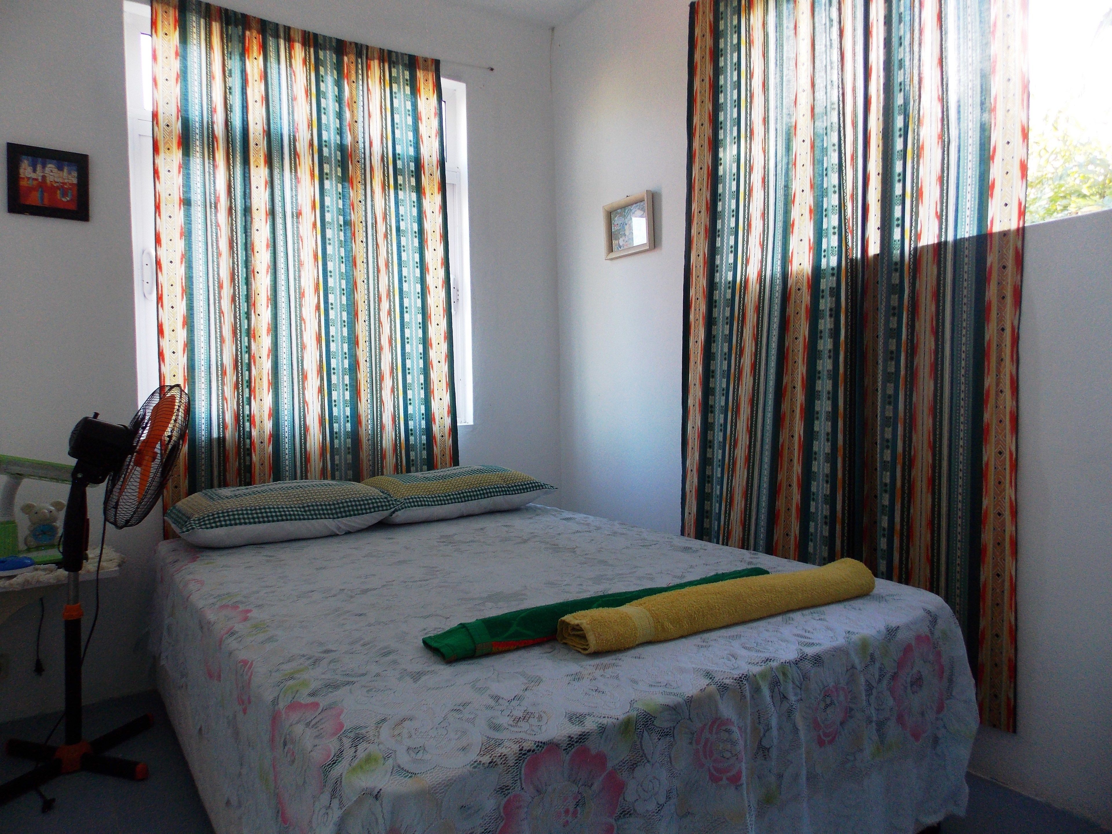 Un dormitorio con cama, almohadas, un ventilador de pie y cortinas.