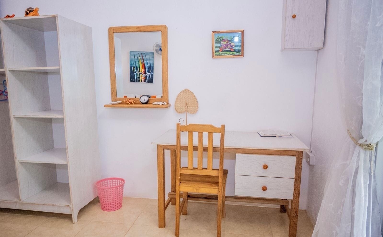 Una habitación con un escritorio de madera, una silla, una estantería, un pequeño estante con un reloj y objetos decorativos, y una cesta rosa en el suelo.