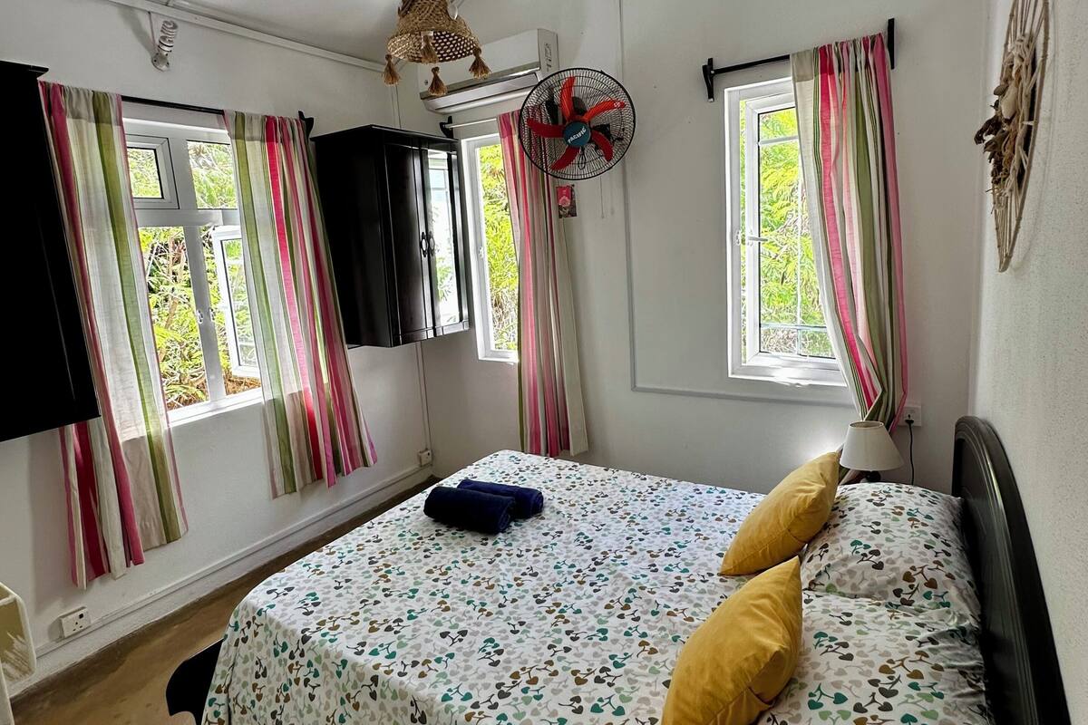 Ein Schlafzimmer mit einem Bett, zwei mit Vorhängen versehenen Fenstern, einem Standventilator und einem Wanddekor.