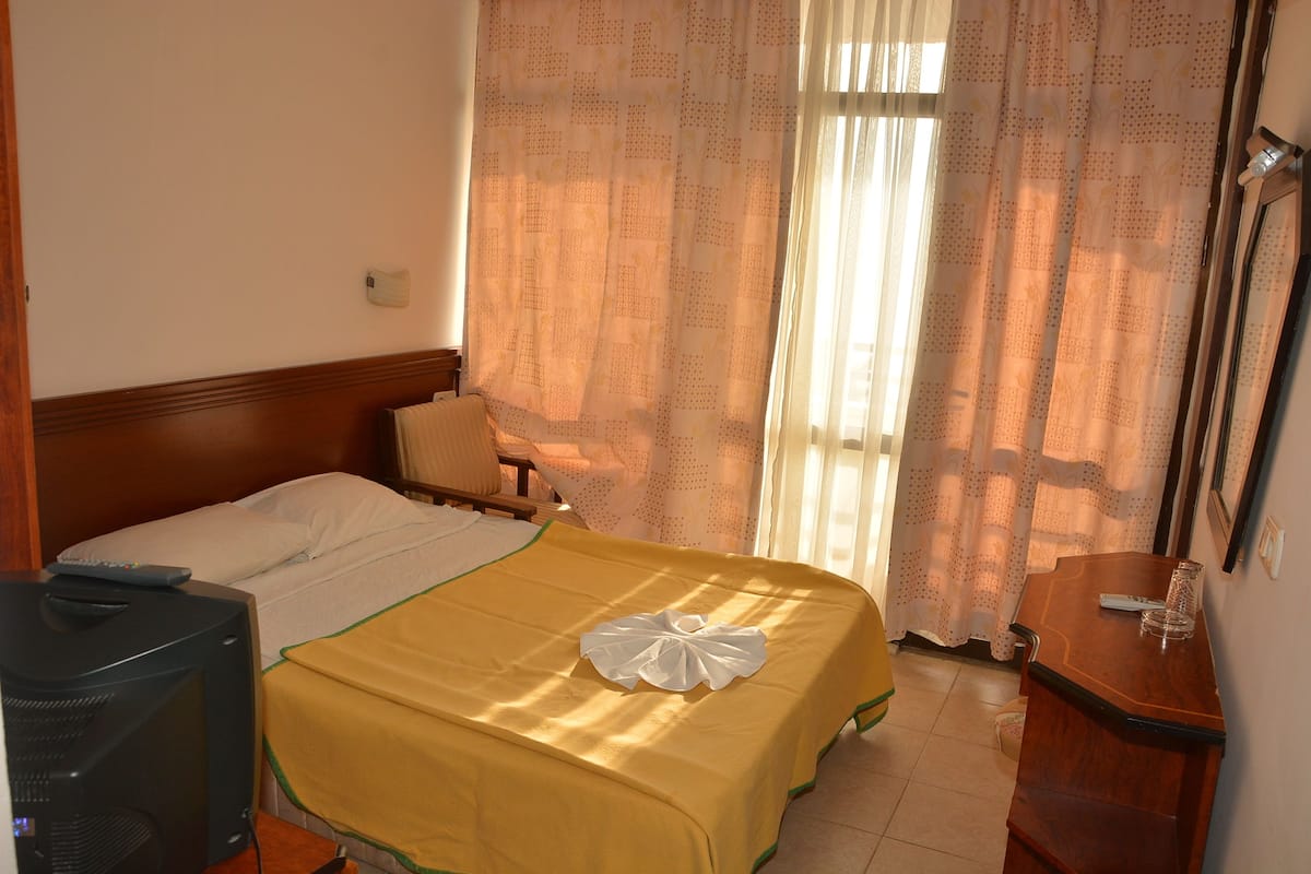 A hotel room with a bed, a television, a wooden nightstand, and a window with curtains.