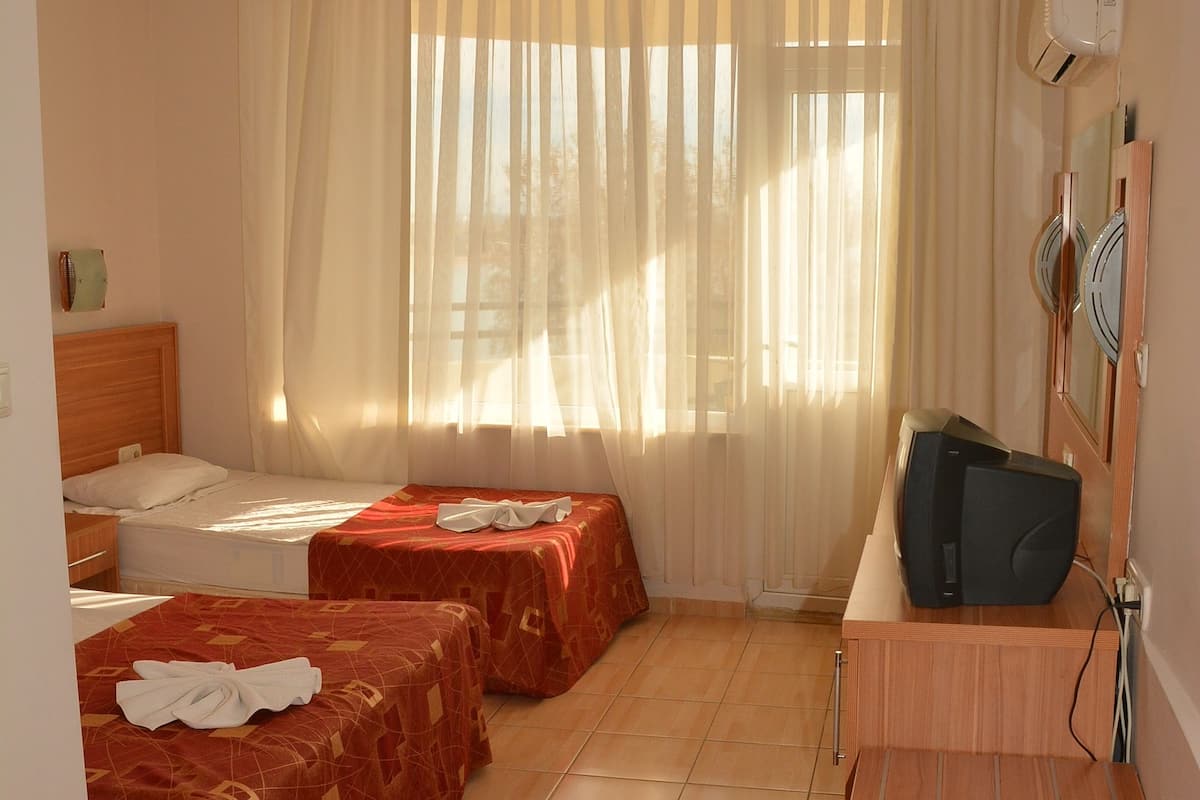 A hotel room with two beds, a wooden desk, a television, and a window with sheer curtains.