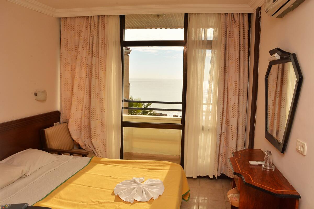 A hotel room with a bed, a wooden desk, a mirror, and a view of the sea through a large window.