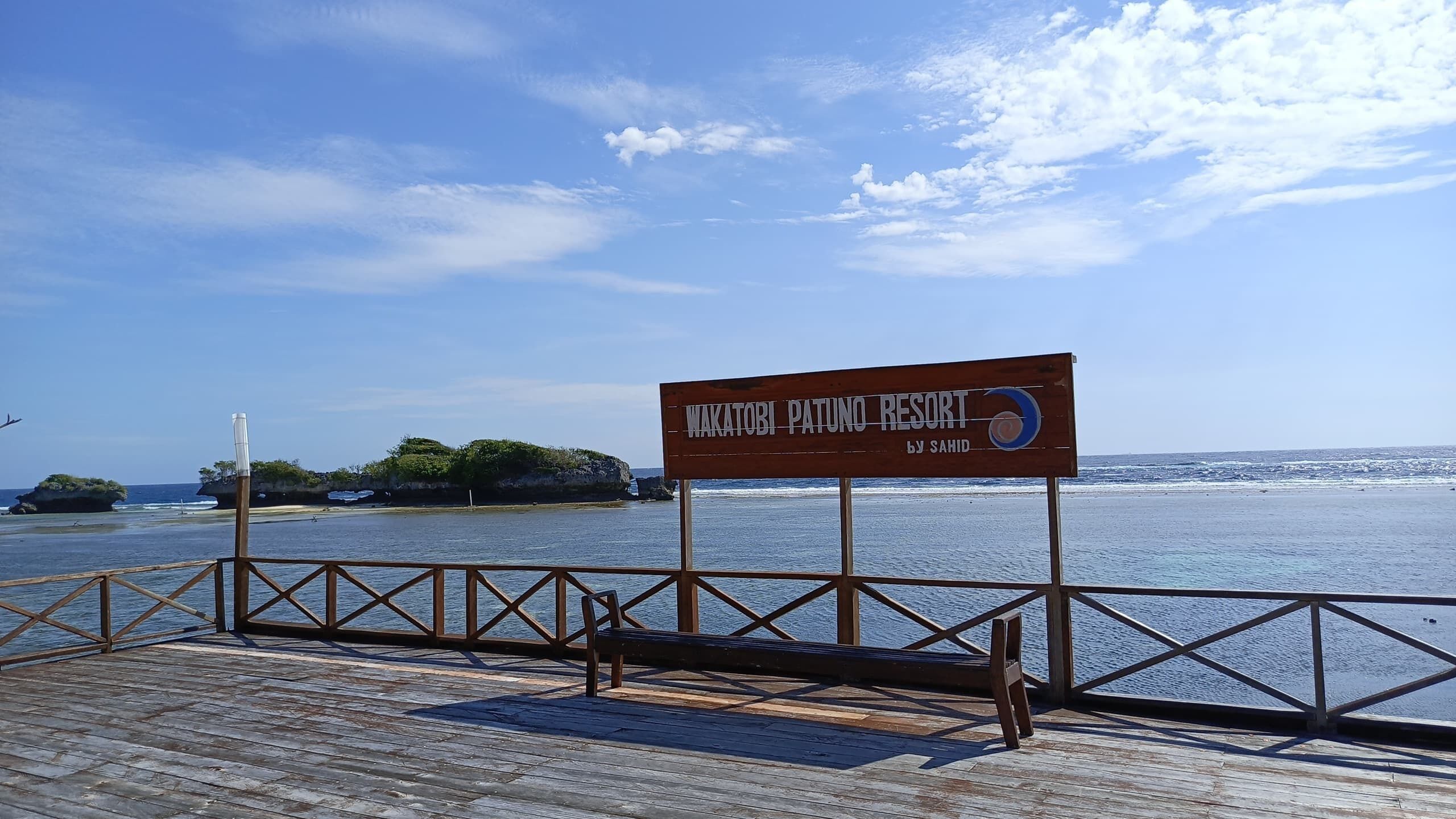 A wooden signboard with the text "Wakatobi Patuno Resort by Sahid" against a backdrop of a clear blue sky and calm sea.