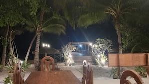 Un espace de restauration en plein air avec une table et des chaises en bois, entouré de palmiers et de plantes en pot, qui mène à un escalier et à une structure illuminée.