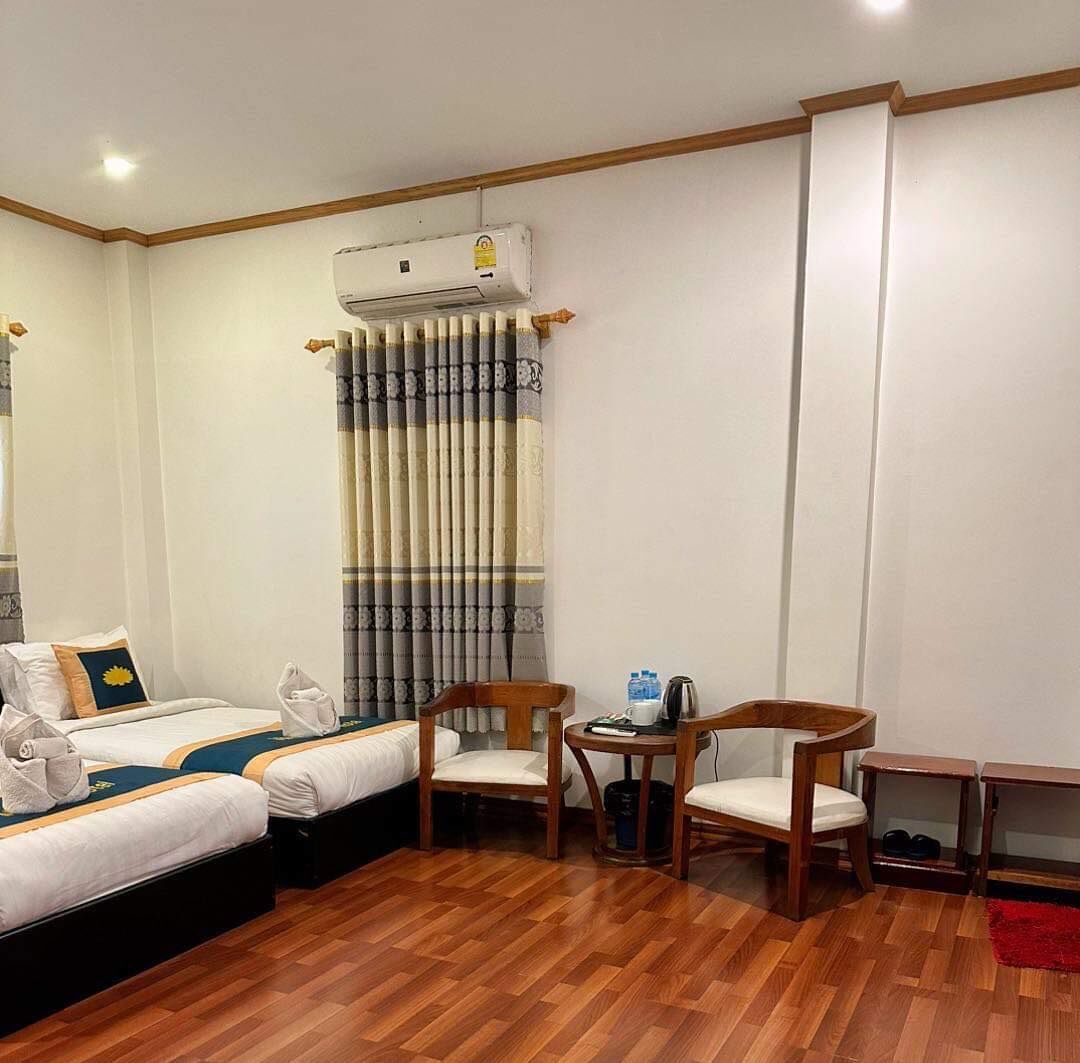 A hotel room with a wooden floor, two beds with white bedding, a small table with a kettle, two chairs, and a red rug.