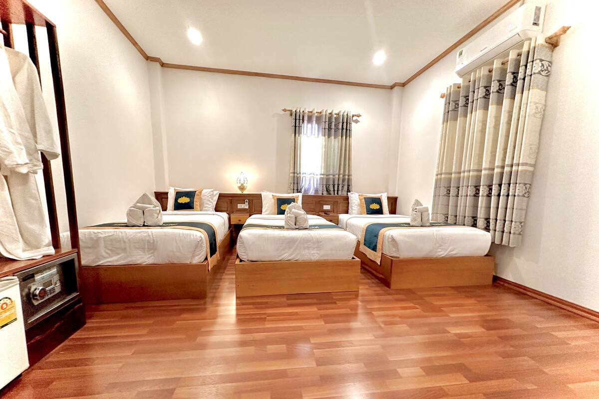 Een hotelkamer met vier eenpersoonsbedden, houten vloeren, een tafeltje met een lamp en een raam met gordijnen.