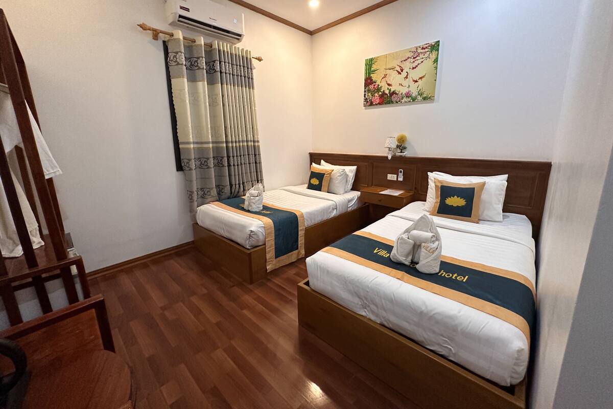 Een hotelkamer met twee bedden, houten vloer, een airconditioningapparaat, een schilderij aan de muur en een stoel.