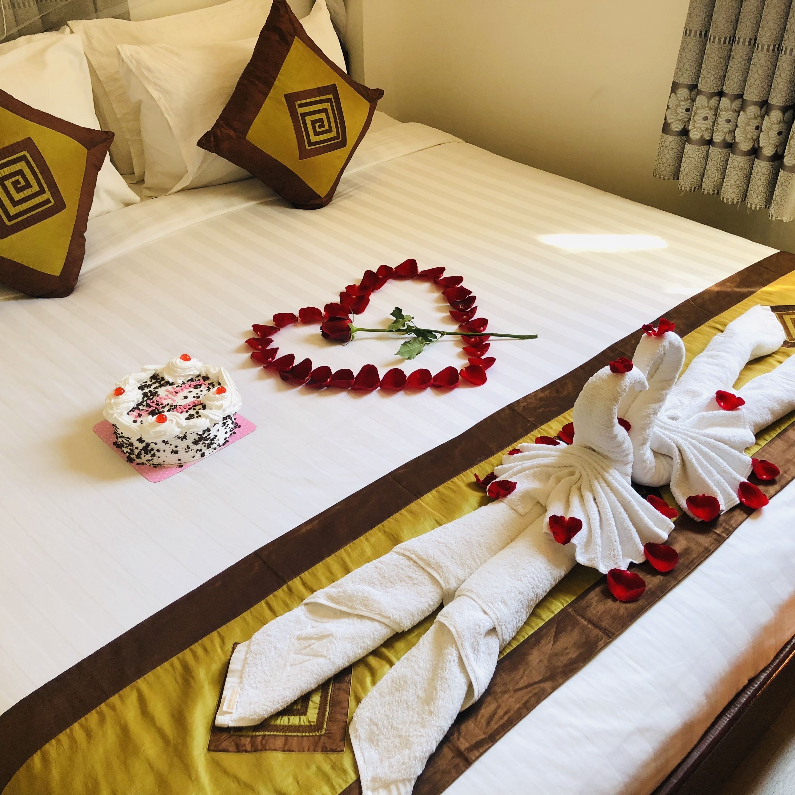 A neatly made bed with a white comforter, a heart-shaped arrangement of red rose petals, a small cake, and a towel arranged in a swan shape.