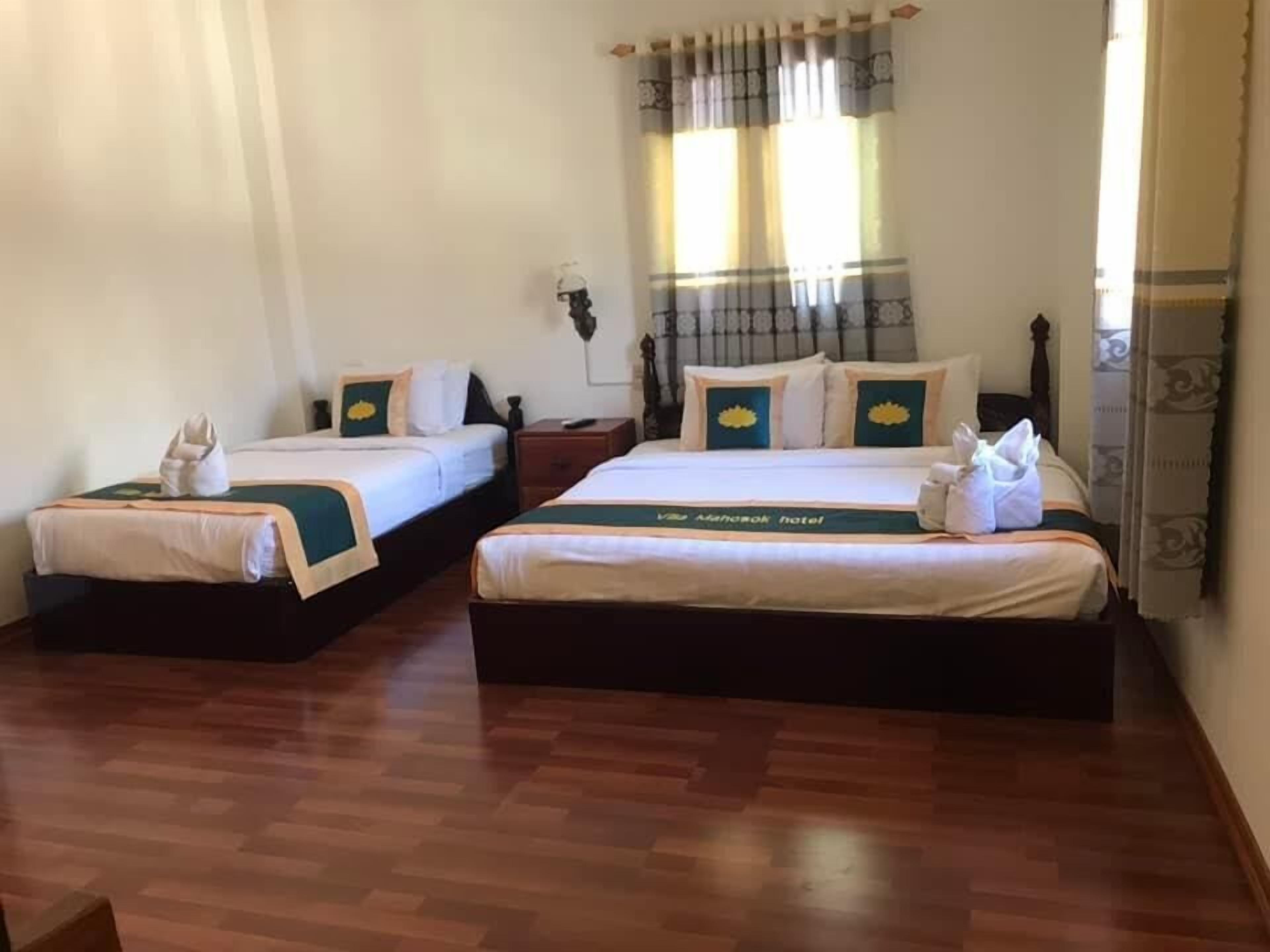 A hotel room with two beds, each with a green bedspread and white linens. There are towels on the beds, and a nightstand with a lamp between the beds.