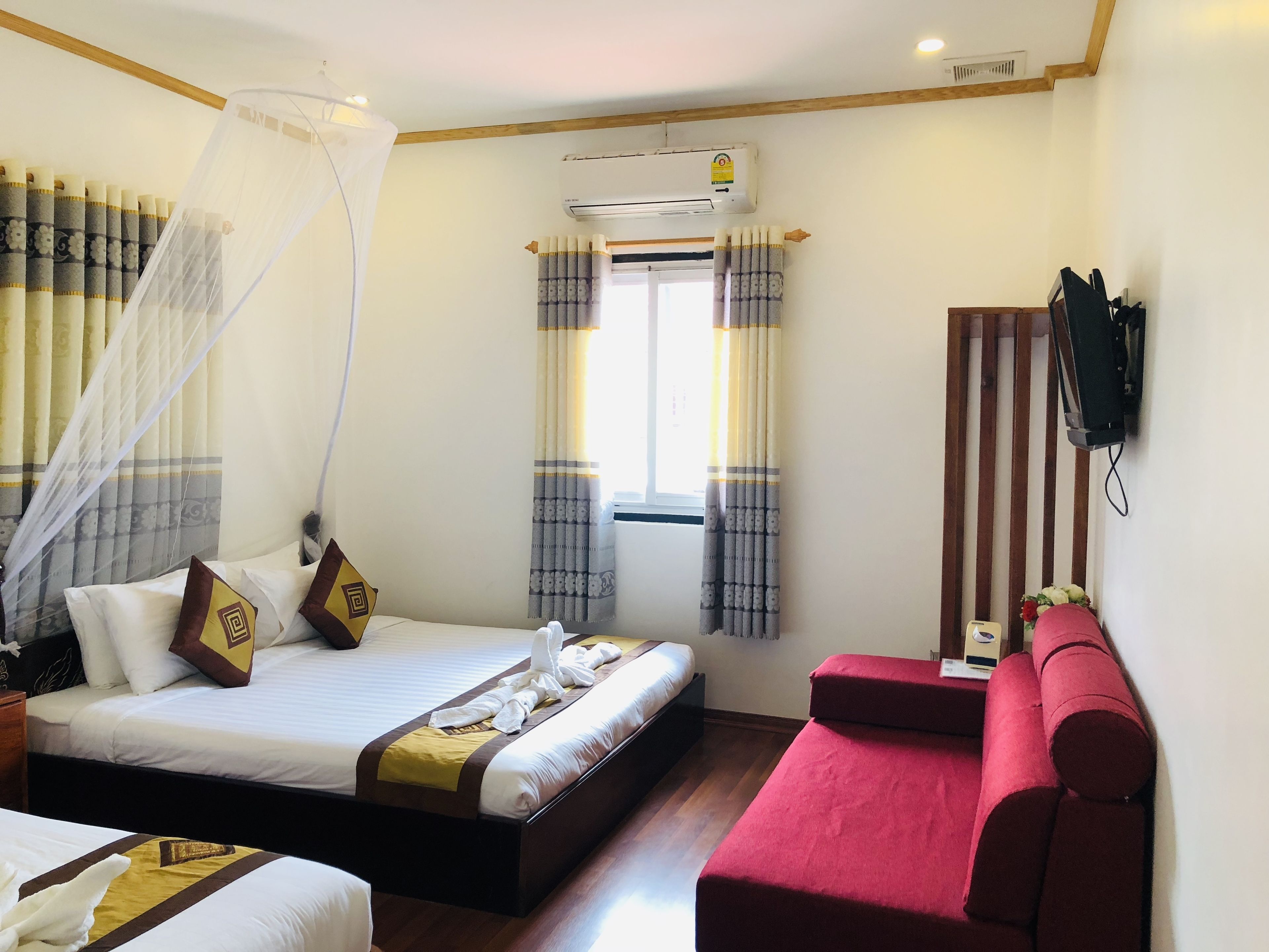 A hotel room with a bed, a red sofa, a TV mounted on the wall, and an air conditioning unit.