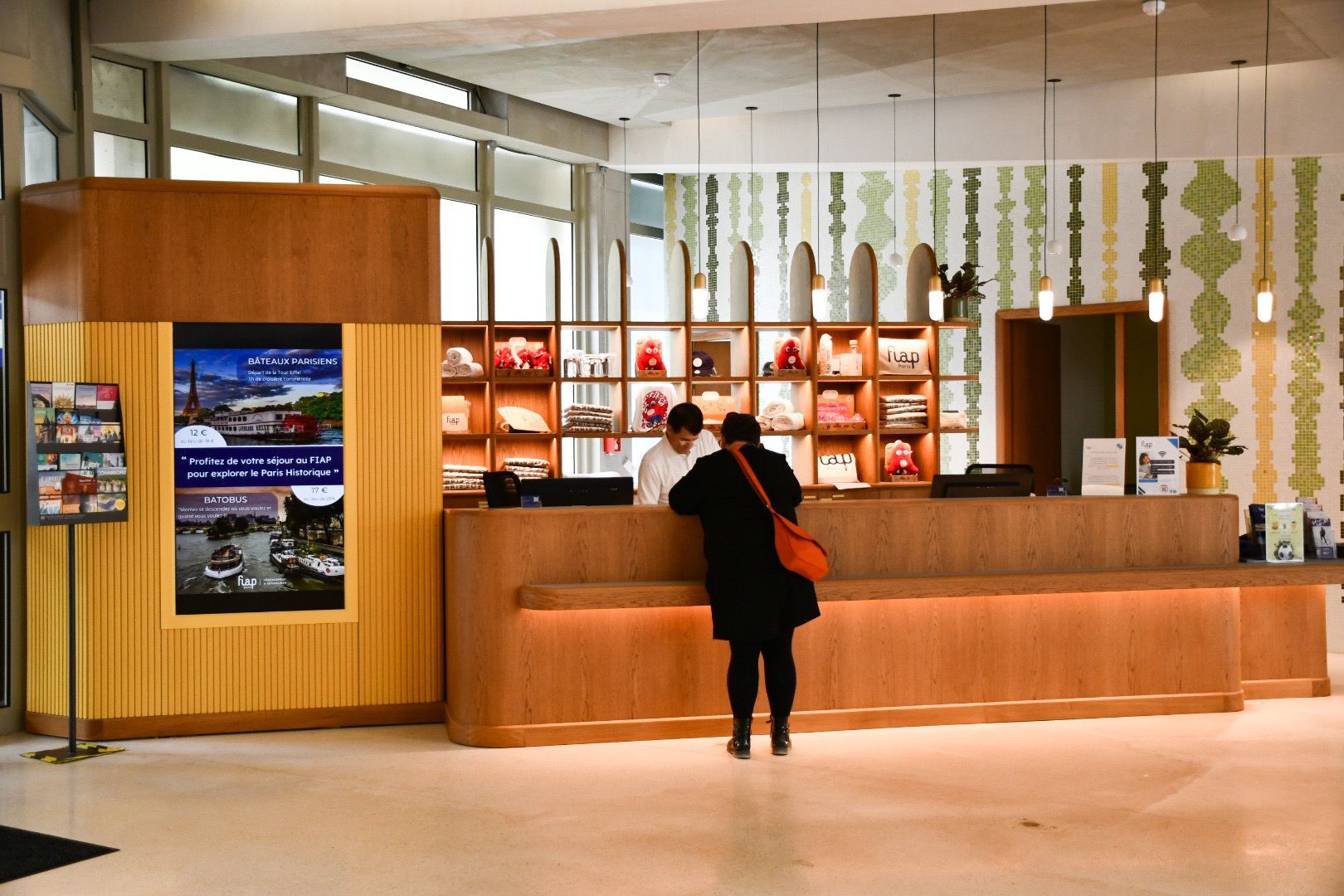 A hotel reception area with a wooden reception desk, a poster advertising Parisian bistros, and a patterned wall behind the desk.
