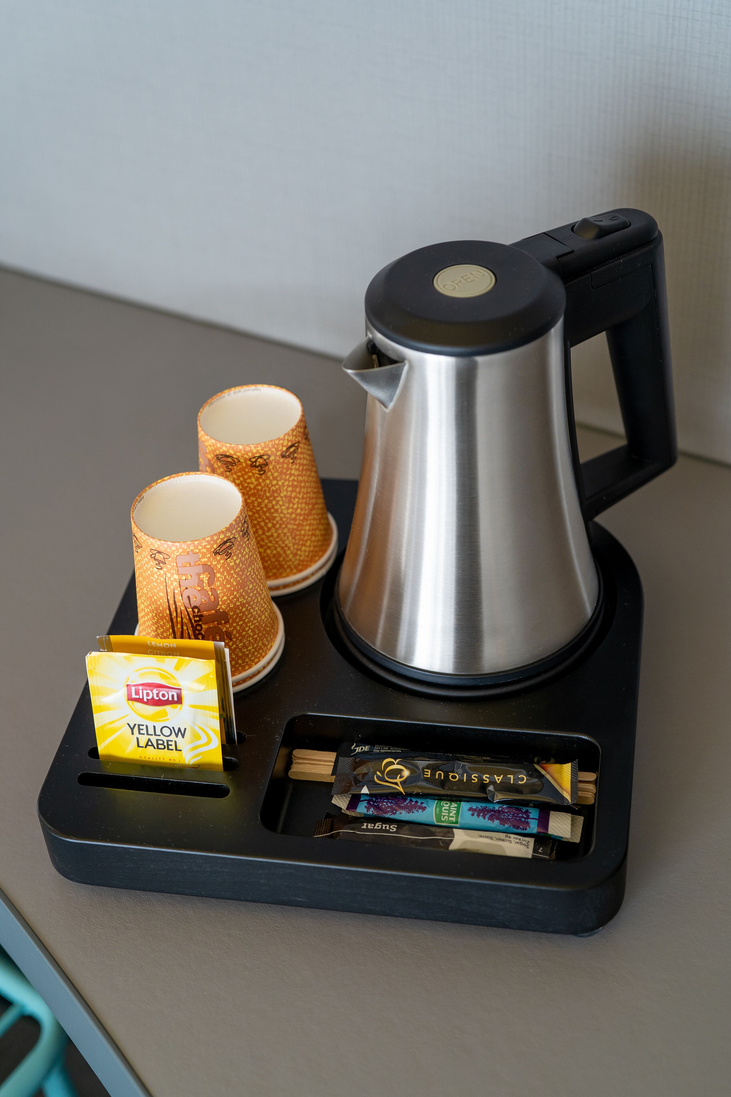 Eine Kaffeemaschine, zwei Pappbecher und eine Packung Lipton Tee auf einem schwarzen Tablett.