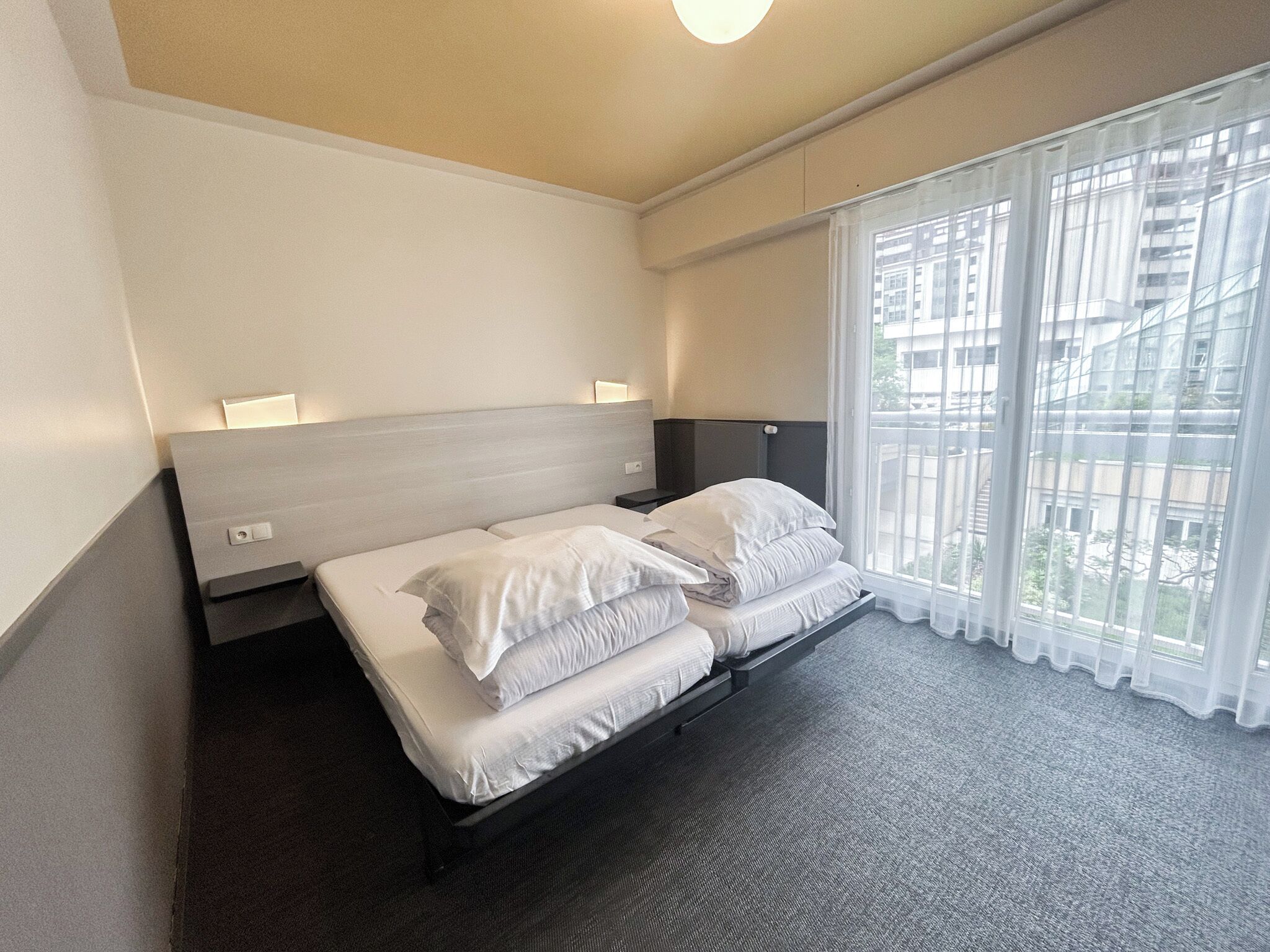 Une chambre d’hôtel avec un lit, une table de chevet, une lampe et une vue sur les immeubles par la fenêtre.