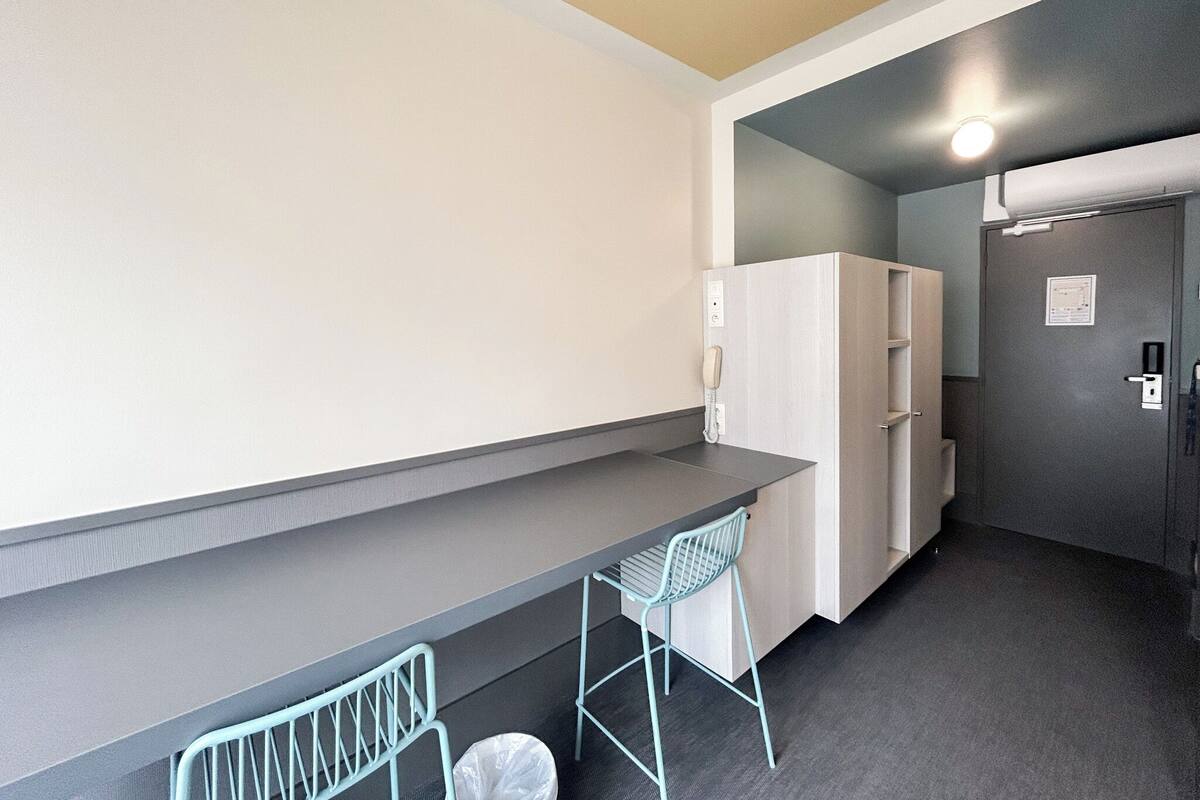 A small kitchenette with a counter, a fridge, and a sink. There is a chair and a trash bin.
