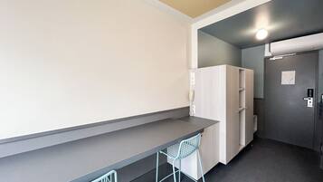 A small kitchenette with a counter, a fridge, and a sink. There is a chair and a trash bin.