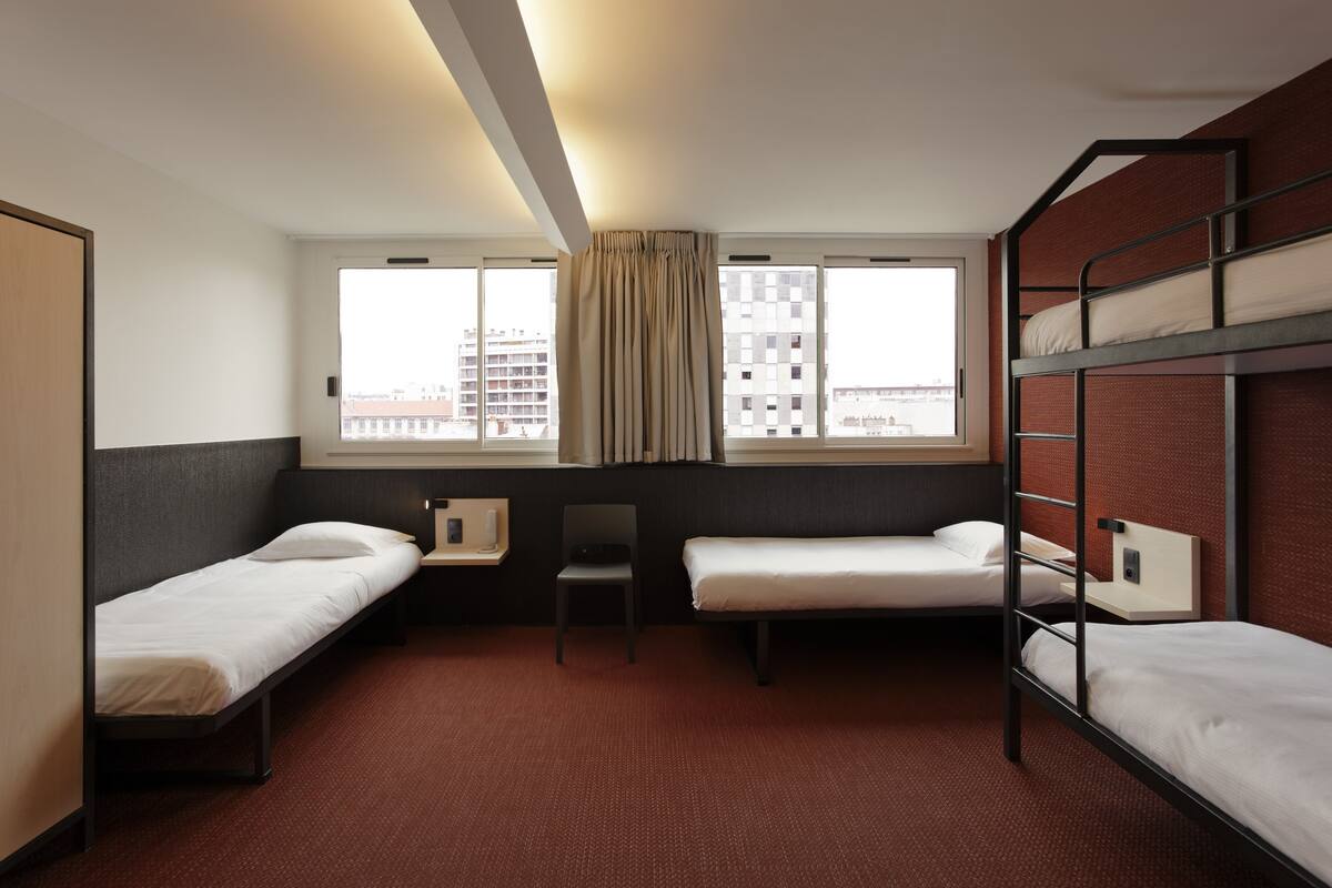 A hotel room with two single beds, a desk, a chair, and a window with curtains.