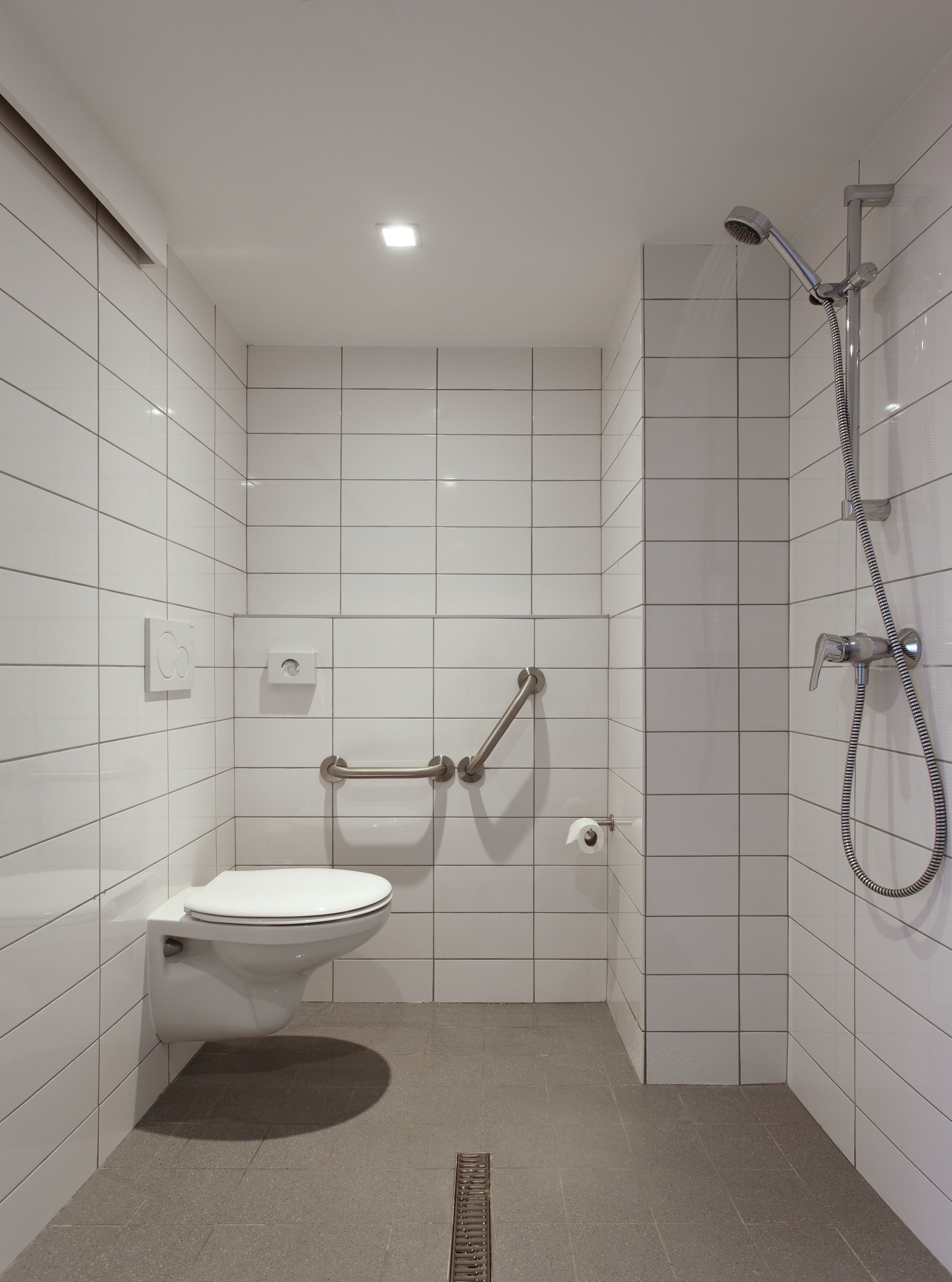 Un baño pequeño y moderno con azulejos blancos, un inodoro, ducha con cabezal de ducha de mano y una barra de apoyo.