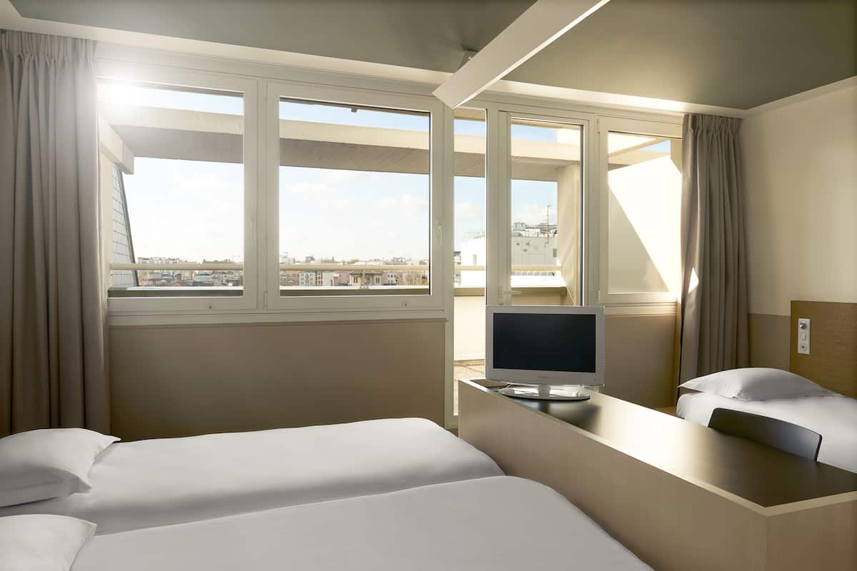 A hotel room with two beds, a desk with a TV, and a large window offering a city view.
