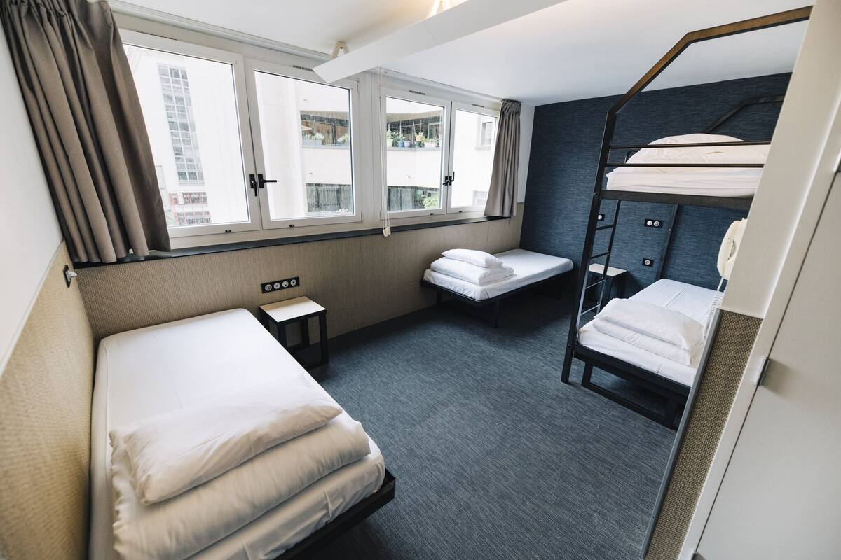 A hotel room with a bunk bed, a single bed, a small table, and a window with curtains.