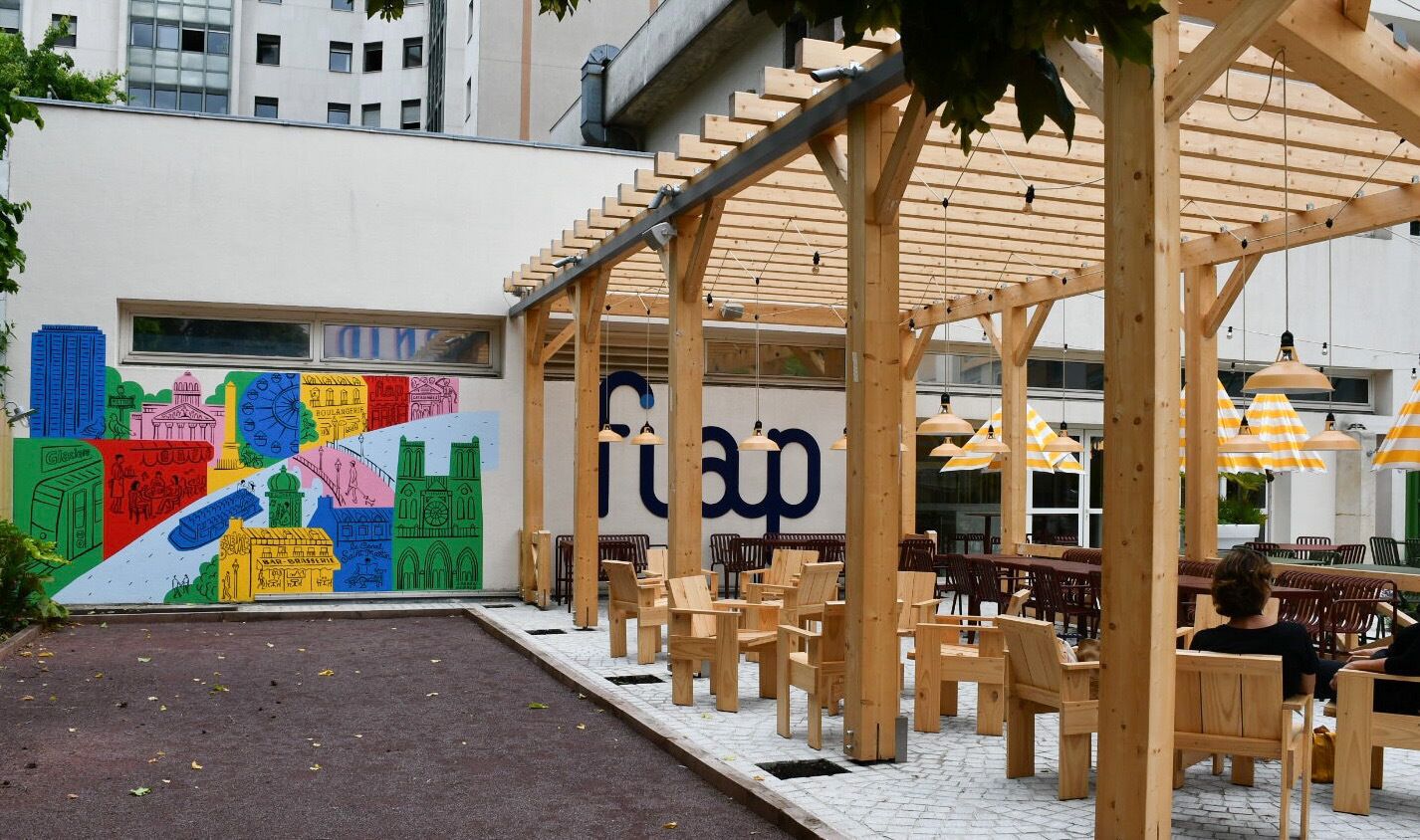 Un espace extérieur aménagé avec des tables et des chaises en bois sous une pergola, une fresque colorée sur le mur adjacent et un bâtiment dont l’entrée est bien visible.