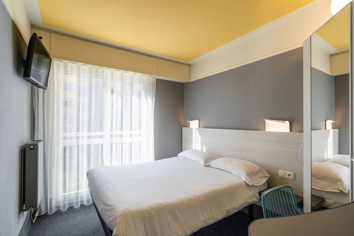 A hotel room with a large bed, a TV mounted on the wall, a mirror, and a window with sheer curtains.