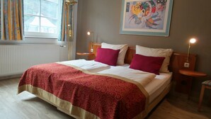 Habitación de hotel con una cama grande, dos mesitas de noche, una ventana con cortinas, un cuadro en la pared y un radiador.