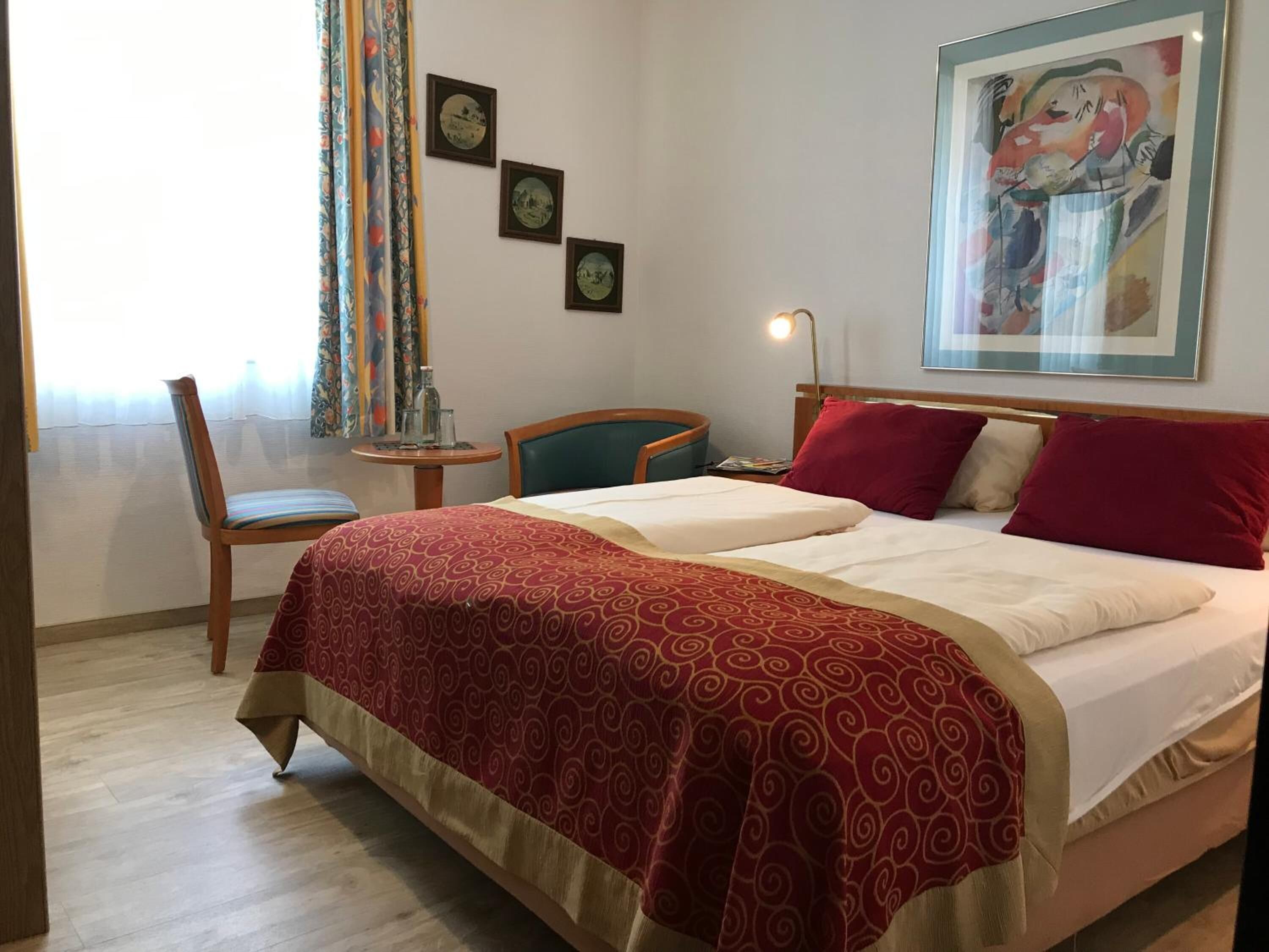 Habitación de hotel con una cama, dos almohadas, una silla, una mesita con una lámpara y un cuadro en la pared.
