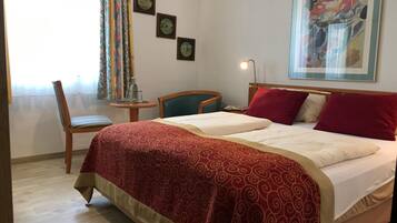 Habitación de hotel con una cama, dos almohadas, una silla, una mesita con una lámpara y un cuadro en la pared.